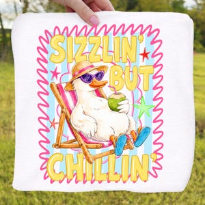 May include: White square towel with a cartoon duck wearing sunglasses and a straw hat, relaxing in a beach chair. The text "SIZZLIN' BUT CHILLIN'" is displayed in yellow. The background has blue stripes and pink accents.