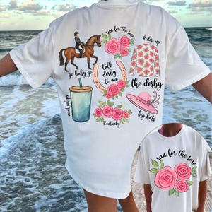 以下が含まれることがあります： ピンクの花柄と「run for the rose」、「go baby go」、「talk derby to me」、「the derby」、「riders up」、「mint juleps」、「Kentucky」、「big hats」という文字が書かれた白いTシャツ。
