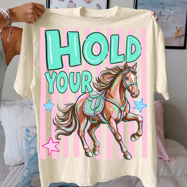 May include: Cream-colored t-shirt featuring a horse illustration with the text "HOLD YOUR" in teal and pink. The horse is brown with a teal saddle and is set against a pink and white striped background. Blue and pink stars are also included.