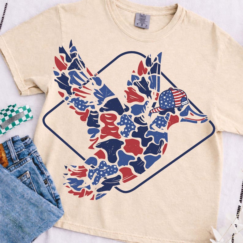 May include: Cream-colored t-shirt featuring a duck design with red, white, and blue camouflage-style patterns. The duck wears a baseball cap with a similar patriotic design. The graphic is enclosed in a dark blue diamond shape. Casual wear.