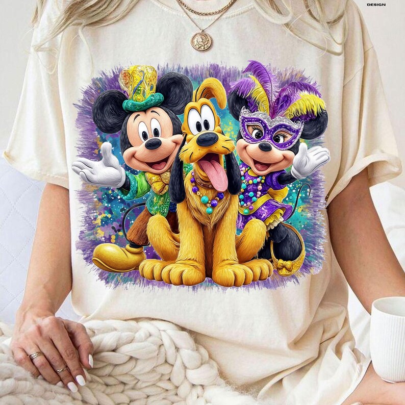 K&ouml;nnte beinhalten: Cremefarbenes T-Shirt mit einem farbenfrohen Aufdruck von Mickey Mouse, Pluto und Minnie Mouse in festlicher Kleidung. Mickey tr&auml;gt ein gr&uuml;nes und goldenes Outfit, Pluto hat eine Halskette und Minnie tr&auml;gt eine lila Maske und ein Kleid. Das Design hat einen Aquarelleffekt.