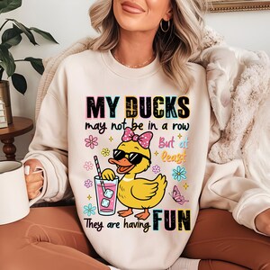 May include: Cream-colored sweatshirt with the text "MY DUCKS may not be in a row But at least They are having FUN." The graphic features a yellow duck wearing sunglasses and a pink bow, holding a drink with ice cubes.