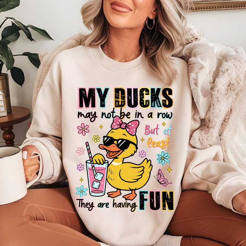 May include: Cream-colored sweatshirt with the text "MY DUCKS may not be in a row But at least They are having FUN." The graphic features a yellow duck wearing sunglasses and a pink bow, holding a drink with ice cubes.
