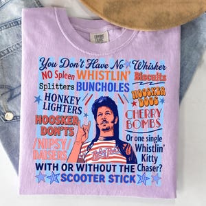 Pu&ograve; includere: T-shirt lilla chiaro con grafica di un uomo e testo. Il testo include frasi come "Whistlin' Biscuits" e "Cherry Bombs". La maglietta &egrave; piegata su una superficie azzurra, con un cappello marrone visibile.