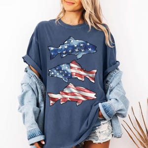 May include: A navy blue t-shirt with three fish designs. The top fish has a blue and white star pattern, the middle fish has a star and stripe pattern, and the bottom fish has a red and white stripe pattern. The shirt has rolled-up sleeves.