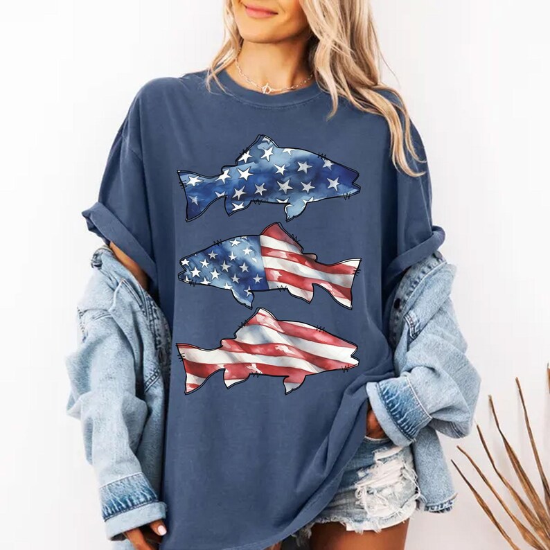 May include: A navy blue t-shirt with three fish designs. The top fish has a blue and white star pattern, the middle fish has a star and stripe pattern, and the bottom fish has a red and white stripe pattern. The shirt has rolled-up sleeves.