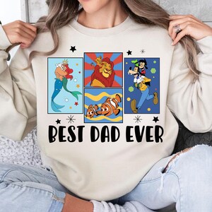 Pu&ograve; includere: Felpa color crema con un design grafico con personaggi dei cartoni animati Disney. Il design include Ariel, Simba, Nemo e Pippo, con la scritta "Best Dad Ever" in nero.