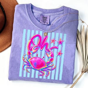May include: A purple Comfort Colors t-shirt featuring a crab graphic with the word "Oh" in pink and blue. The design includes blue vertical stripes and pink stars. A gold necklace is draped across the shirt.
