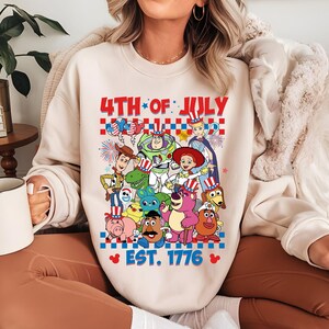 May include: A cream-colored sweatshirt with a graphic of Toy Story characters wearing red, white, and blue patriotic hats. The text "4th of July Est. 1776" is printed on the sweatshirt.