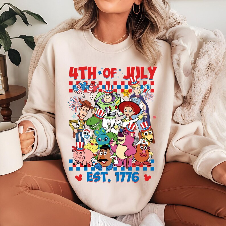 May include: A cream-colored sweatshirt with a graphic of Toy Story characters wearing red, white, and blue patriotic hats. The text "4th of July Est. 1776" is printed on the sweatshirt.