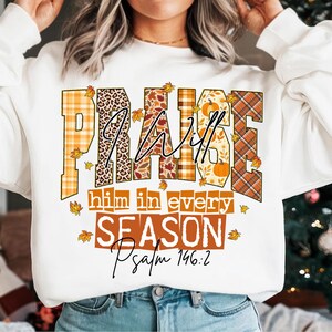 May include: White sweatshirt with the words "PRAISE HIM in every SEASON Psalm 146:2" in a fall-themed design. The word "PRAISE" is in orange plaid, leopard print, and floral patterns. The text is surrounded by falling leaves.