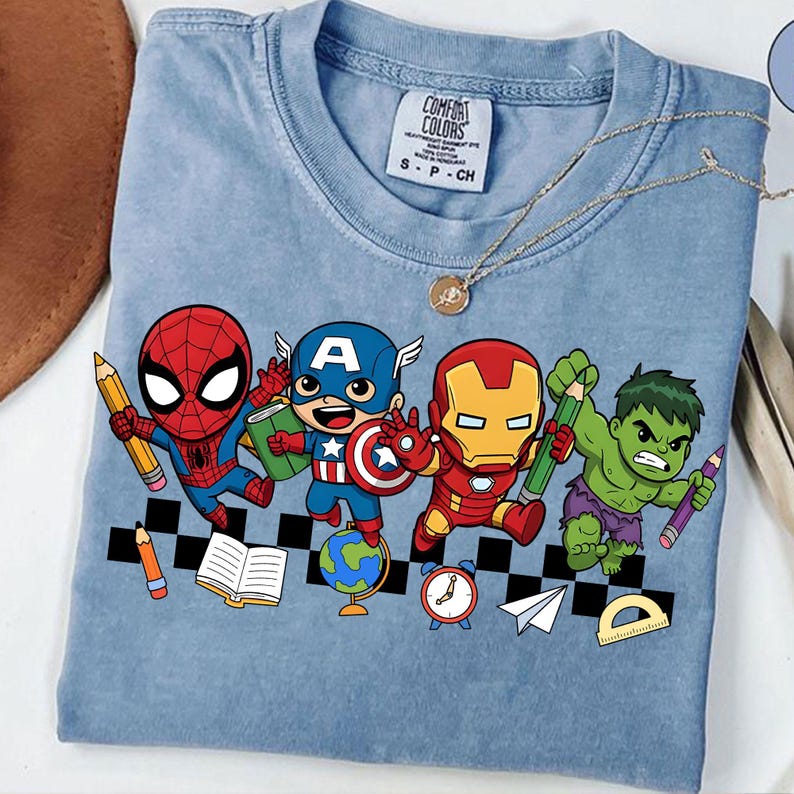 May include: Light blue t-shirt featuring cartoon versions of Spider-Man, Captain America, Iron Man, and the Hulk, each holding school supplies. The shirt includes the text "COMFORT COLORS" and size information.