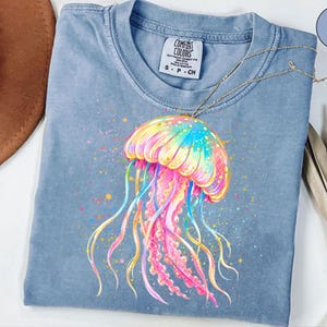 May include: A light blue Comfort Colors t-shirt featuring a colorful jellyfish design. The jellyfish is depicted with vibrant hues of pink, yellow, blue, and green, with long, flowing tentacles. The shirt has a classic crew neck and short sleeves.