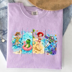 May include: A light purple Comfort Colors t-shirt featuring a colorful beach scene with characters. The graphic includes a man in a Hawaiian shirt, a green dinosaur on a pink flamingo float, a girl in a red swimsuit, and Buzz Lightyear.