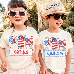 May include: Two children wearing cream-coloured t-shirts with patriotic designs. The shirts feature an American flag popsicle, balloons, and an American flag. One shirt says "Sophia" and the other says "William". Both children are wearing sunglasses.