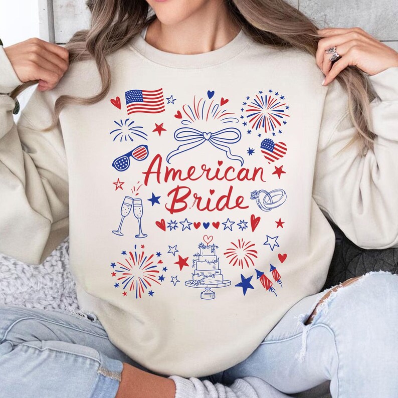 May include: Cream-colored sweatshirt with a patriotic design. The graphic includes an American flag, fireworks, hearts, stars, and the text "American Bride" in red and blue. Features wedding-themed illustrations.
