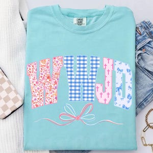 May include: A light blue Comfort Colors t-shirt with the letters "WWJD" in a decorative font. The "W" is floral, the "W" is blue gingham, the "J" is pink with crosses, and the "D" is patterned with blue bows. A pink bow graphic is below.