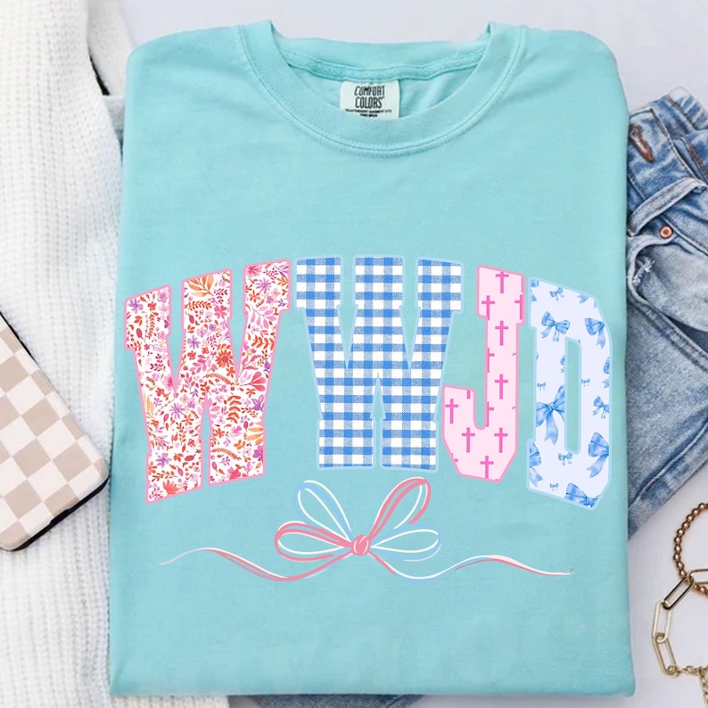 May include: A light blue Comfort Colors t-shirt with the letters "WWJD" in a decorative font. The "W" is floral, the "W" is blue gingham, the "J" is pink with crosses, and the "D" is patterned with blue bows. A pink bow graphic is below.