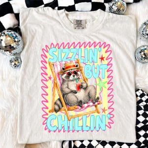 May include: Cream-colored t-shirt featuring a raccoon in sunglasses and a straw hat, relaxing in a beach chair with a drink. The shirt has the text "Sizzlin' But Chillin'" in a colorful design. The shirt is a Comfort Colors brand.