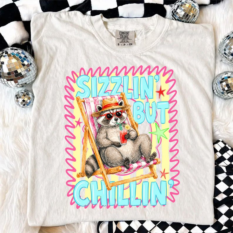 May include: Cream-colored t-shirt featuring a raccoon in sunglasses and a straw hat, relaxing in a beach chair with a drink. The shirt has the text "Sizzlin' But Chillin'" in a colorful design. The shirt is a Comfort Colors brand.