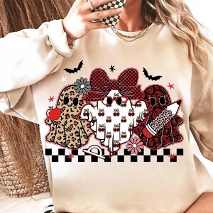 May include: Beige sweatshirt featuring three ghost characters with Halloween and school-themed designs. One ghost has a leopard print, another has a bow, and the third holds a pencil. Bats and stars are also included.