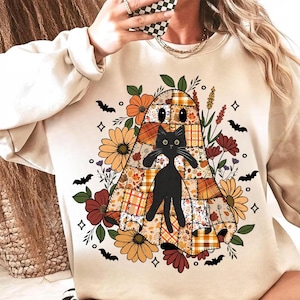 May include: Beige sweatshirt featuring a Halloween-themed design. The graphic includes a patchwork ghost with a black cat, surrounded by flowers, bats, and decorative elements. The design incorporates orange, brown, and black colors.