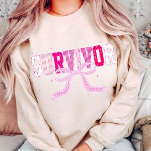 May include: A beige sweatshirt with the word "SURVIVOR" in pink and white block letters. A pink ribbon design is below the word. The sweatshirt has long sleeves and a crew neck.