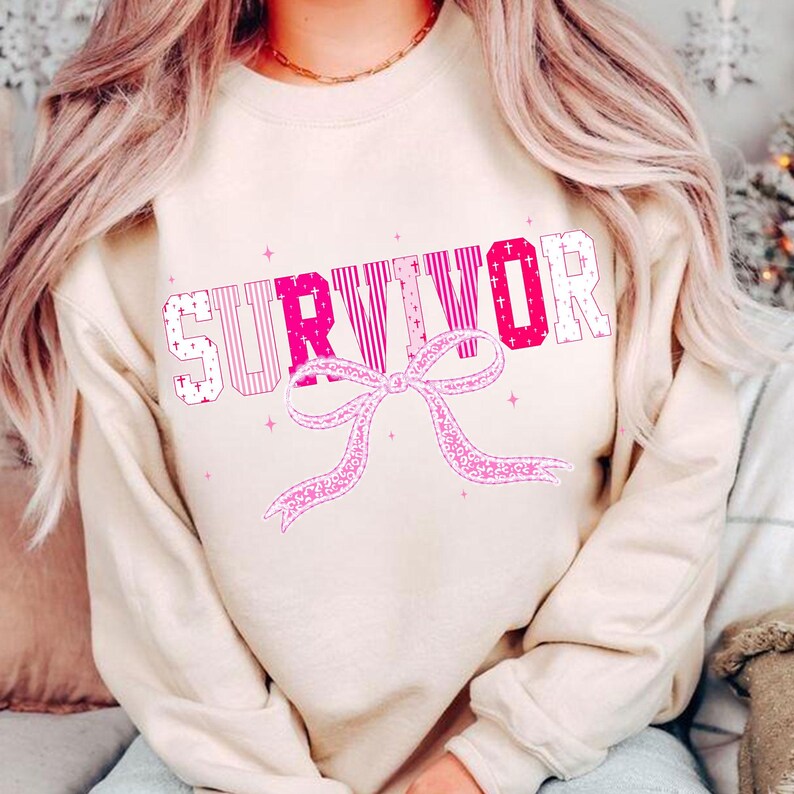 May include: A beige sweatshirt with the word "SURVIVOR" in pink and white block letters. A pink ribbon design is below the word. The sweatshirt has long sleeves and a crew neck.