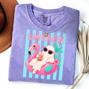K&ouml;nnte beinhalten: Lilafarbenes T-Shirt mit dem Schriftzug "SUMMERWEEN" und einer Cartoon-Geistfigur mit Sonnenbrille, die auf einem rosa Flamingo-Schwimmer entspannt. Das Design umfasst eine Palme, Blumen und blaue vertikale Streifen. Marke Comfort Colors.