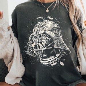 May include: Dark gray sweatshirt featuring a detailed black and white illustration of Darth Vader's helmet. The design includes space ships and a starry background, creating a galactic theme. The sweatshirt is a casual wear item.