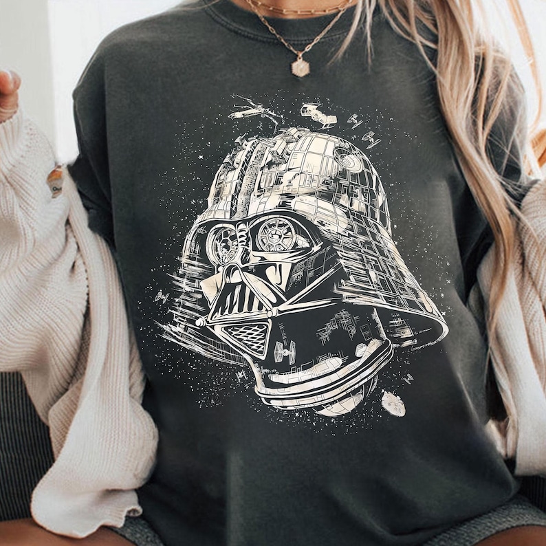 May include: Dark gray sweatshirt featuring a detailed black and white illustration of Darth Vader's helmet. The design includes space ships and a starry background, creating a galactic theme. The sweatshirt is a casual wear item.