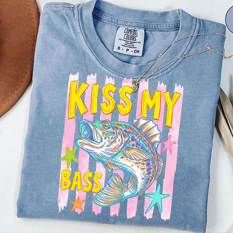 May include: A blue Comfort Colors t-shirt with the phrase "KISS MY BASS" in yellow letters. The shirt features a colorful illustration of a fish, pink stripes, and star graphics. A gold necklace is draped across the shirt.