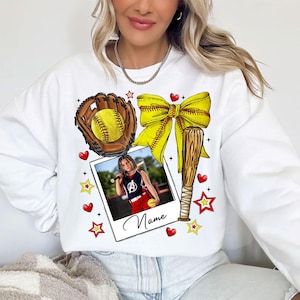 May include: White sweatshirt with a softball theme. It features a baseball glove, bat, ball, bow, and stars. A photo of a girl playing softball is in the center of the design. The text "Name" is below the photo.