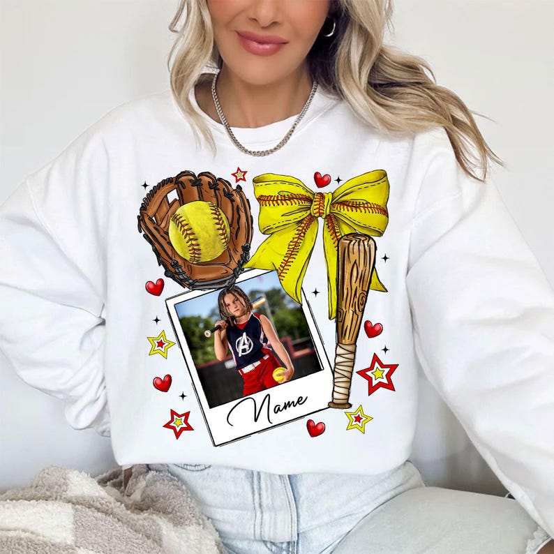 May include: White sweatshirt with a softball theme. It features a baseball glove, bat, ball, bow, and stars. A photo of a girl playing softball is in the center of the design. The text "Name" is below the photo.
