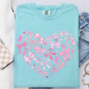 May include: A light blue t-shirt with a heart-shaped design made of pink bows and ribbons. The shirt has the Comfort Colors brand tag. The bows and ribbons are in various styles and patterns, with small pink hearts scattered throughout.