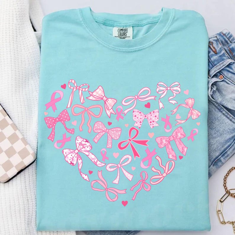 May include: A light blue t-shirt with a heart-shaped design made of pink bows and ribbons. The shirt has the Comfort Colors brand tag. The bows and ribbons are in various styles and patterns, with small pink hearts scattered throughout.