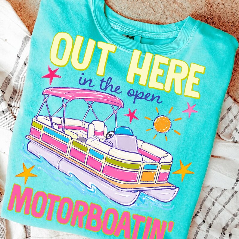 May include: A turquoise crew neck t-shirt with a colorful graphic of a pontoon boat. The text "OUT HERE in the open MOTORBOATIN'" is displayed in yellow and pink. The design includes stars and a sun.