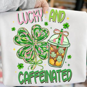 Puede incluir: Sudadera blanca con un diseño del Día de San Patricio. El gráfico incluye un gran trébol, una bebida de café con detalles de trébol y las palabras "LUCKY AND CAFFEINATED" en verde y rosa.
