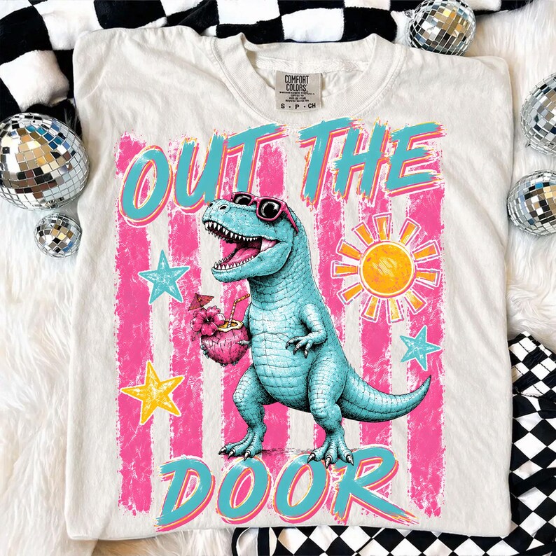 May include: Off-white t-shirt with a turquoise dinosaur wearing sunglasses and holding a tropical drink. The shirt features the words "OUT THE DOOR" in a retro font, with pink and blue accents. A sun and stars complete the design.