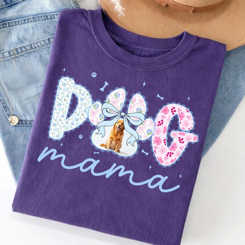 May include: A purple t-shirt with the words "Dog mama" in a light blue script. The word "Dog" is designed with a paw print and floral patterns. A golden retriever is in the center of the paw print, with a light blue bow.