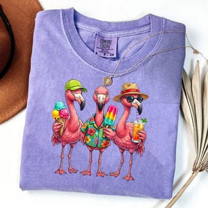 May include: A purple Comfort Colors t-shirt featuring a graphic of three pink flamingos. One flamingo holds an ice cream cone, another a popsicle, and the third a cocktail. The flamingos wear hats and sunglasses, with the text "COMFORT COLORS" on the shirt.