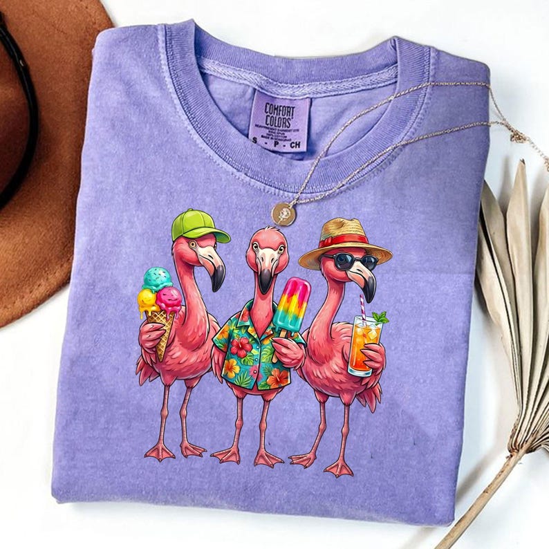 May include: A purple Comfort Colors t-shirt featuring a graphic of three pink flamingos. One flamingo holds an ice cream cone, another a popsicle, and the third a cocktail. The flamingos wear hats and sunglasses, with the text "COMFORT COLORS" on the shirt.