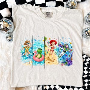 May include: White Comfort Colors t-shirt featuring a colorful graphic of cartoon characters enjoying a beach vacation. The design includes a boy in a Hawaiian shirt, a dinosaur on a flamingo float, a girl in a bikini, and Buzz Lightyear.