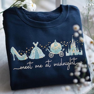 May include: Navy blue sweatshirt featuring a Cinderella-themed design. The graphic includes a glass slipper, ball gown, carriage, and castle, with the text "meet me at midnight" in white script. Gold stars accent the design.