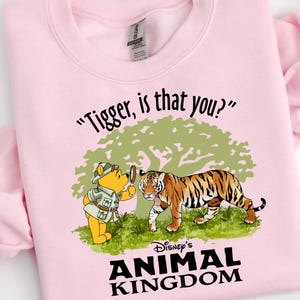 Puede incluir: Sudadera rosa con una imagen de dibujos animados de Winnie de Pooh mirando a un tigre a trav&eacute;s de una lupa. El texto "Tigger, is that you?" est&aacute; por encima de la imagen y "Disney's Animal Kingdom" est&aacute; por debajo de la imagen.
