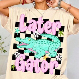 May include: Cream-colored t-shirt featuring a cartoon alligator with the words "Later Gator" in pink. The design includes a black and white checkered background, flowers, and stars.