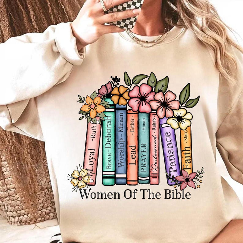 May include: Beige sweatshirt with a colorful graphic of books and flowers. The books are labeled with words like "Loyal", "Brave", and "Faith". The text "Women Of The Bible" is at the bottom.