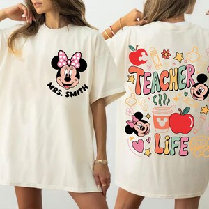 May include: Cream-coloured t-shirt with a Minnie Mouse design and the text "Mrs. Smith" on the front. The back features "Teacher Life" with school-themed graphics, including apples, pencils, and Mickey Mouse.