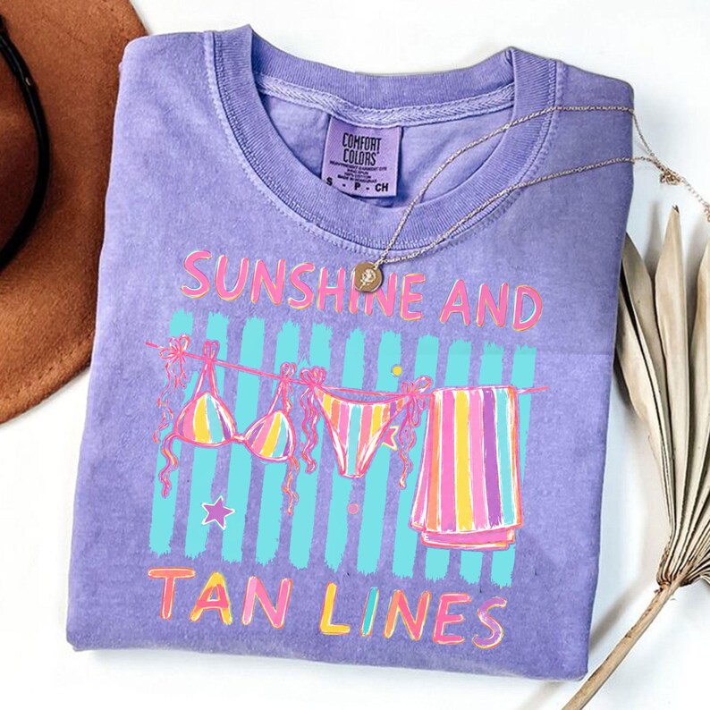 Peut inclure: Un t-shirt violet Comfort Colors avec le texte "Sunshine and Tan Lines" en rose et jaune. Le graphisme comprend un bikini, un bas de bikini et une serviette ray&eacute;e sur une corde &agrave; linge. Un collier dor&eacute; est drap&eacute; sur le t-shirt.