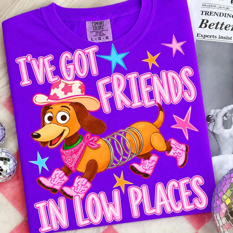May include: A bright purple t-shirt featuring a graphic of a brown dachshund toy with a metal spring body, wearing a pink cowboy hat, a pink bandana, and pink cowboy boots. The text reads I'VE GOT FRIENDS IN LOW PLACES with colorful stars.